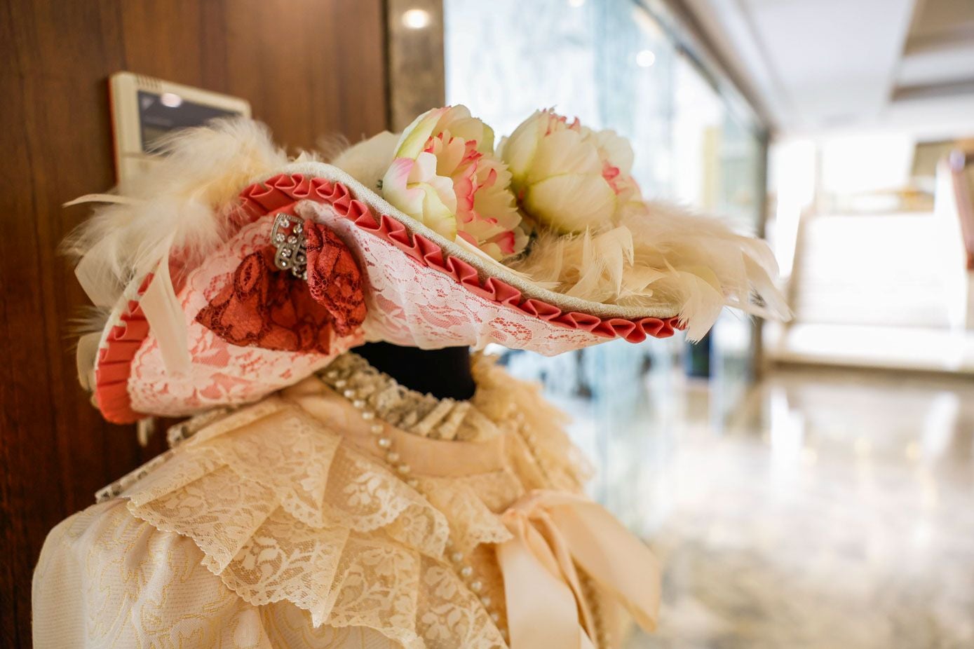 Sombrero de encaje adornado con un plisado que recorre la base del accesorio, además de plumas, flores artificiales y un broche brillante.
