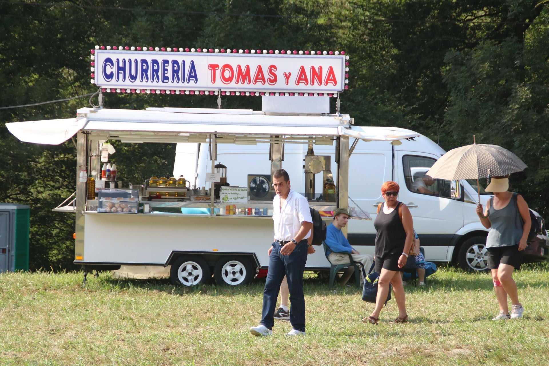 Unos asistentes caminan protegiéndose del sol ante una churrería.