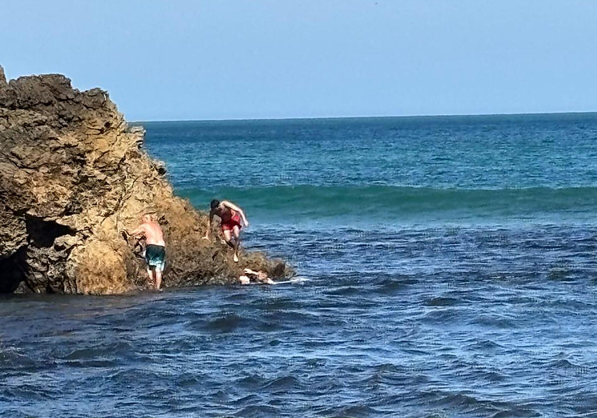 El hombre se agarra a la roca, mientras en el agua el ertzaintza sujeta a la mujer exhausta y el hombre del bañador rojo se acerca para ayudarlos.