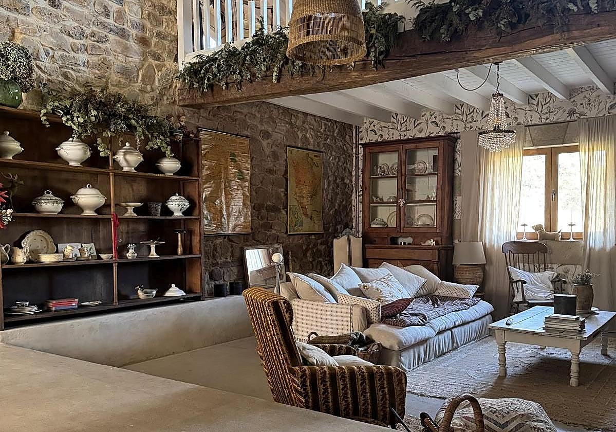 El salón de La Madriguera de Cristina tiene las paredes de piedra, las vigas de madera y los techos blancos. Las estanterías llenas de vajillas antiguas y el papel pintado colocado estratégicamente confieren calidez al espacio.