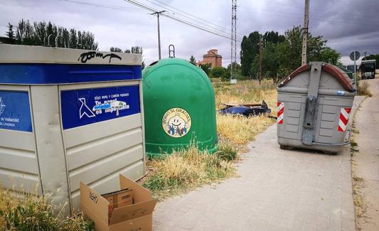 Contenedores entre los hierbajos secos y un parachoques abandonado al fondo. 