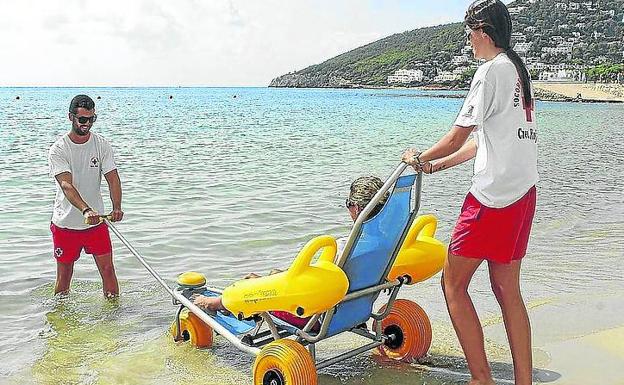 Dos miembros de la Cruz Roja ayudan a una persona con discapacidad a bañarse en una playa. 