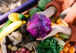 Una mujer sostiene una col lombarda sobre una caja llena de frutas y verduras.