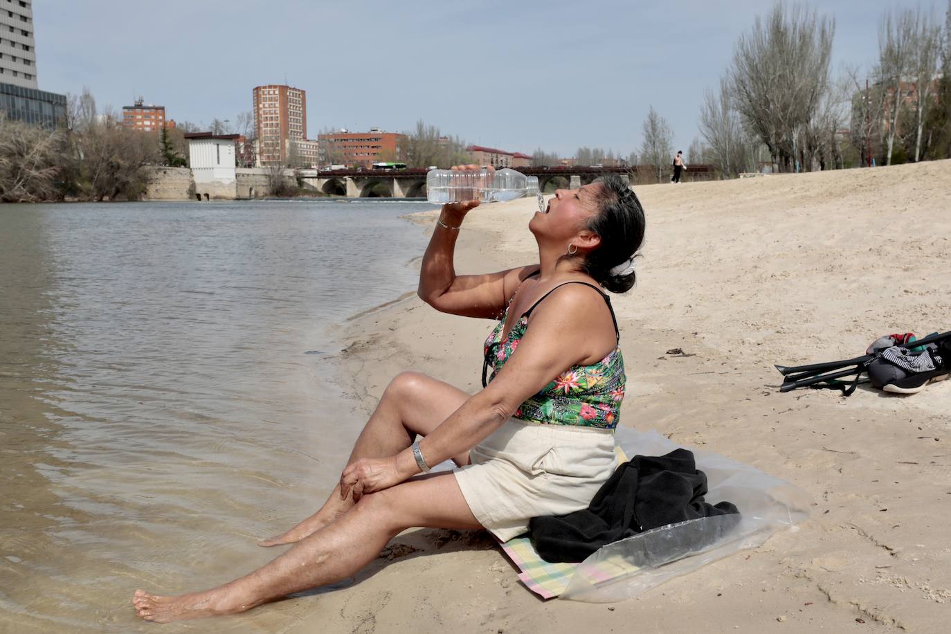 Una señora bebe agua y mete los pies en el río.