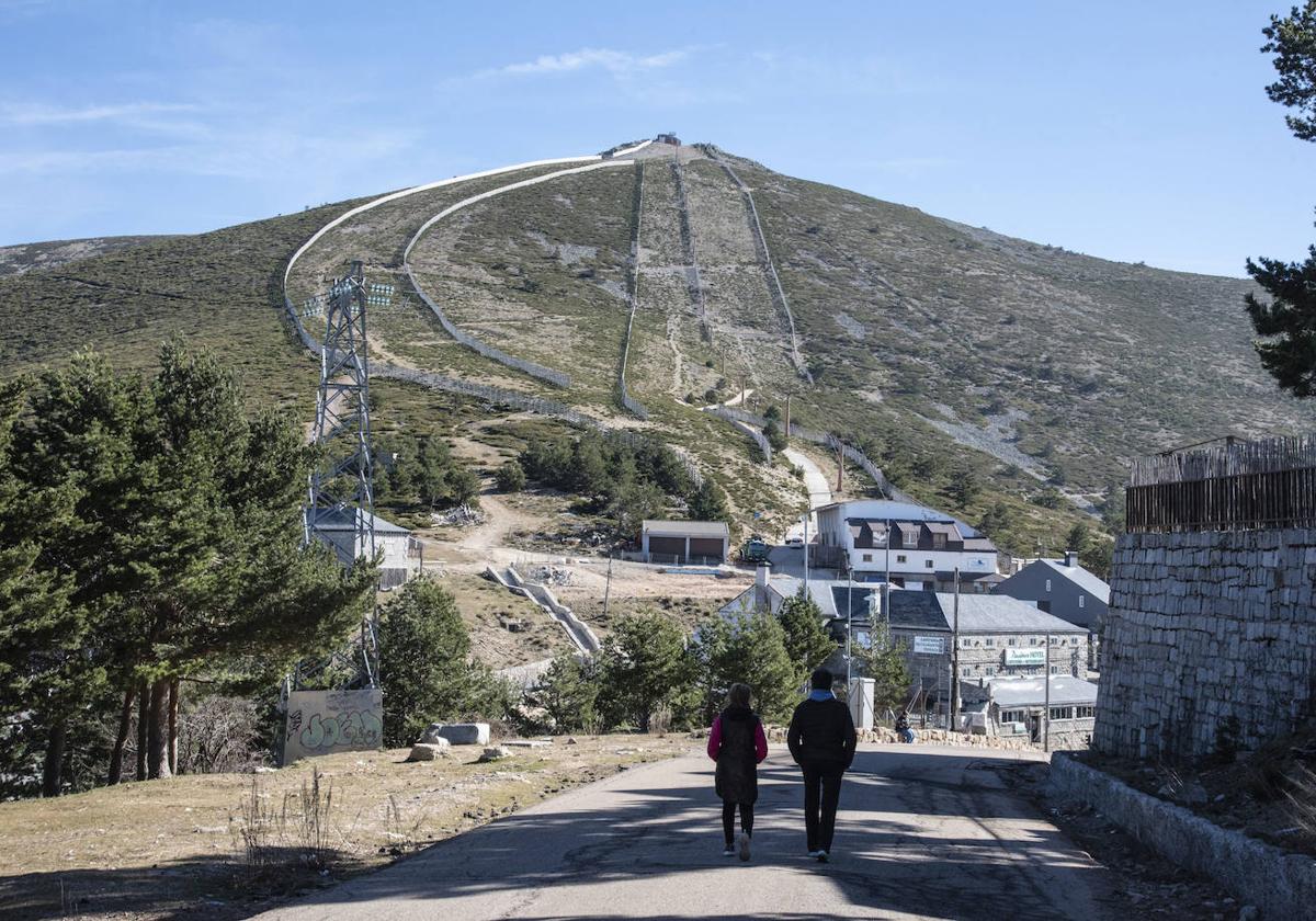 Dos personas transitan junto a la estación de esquí de Navacerrada, con la pista Guarramillas al fondo.