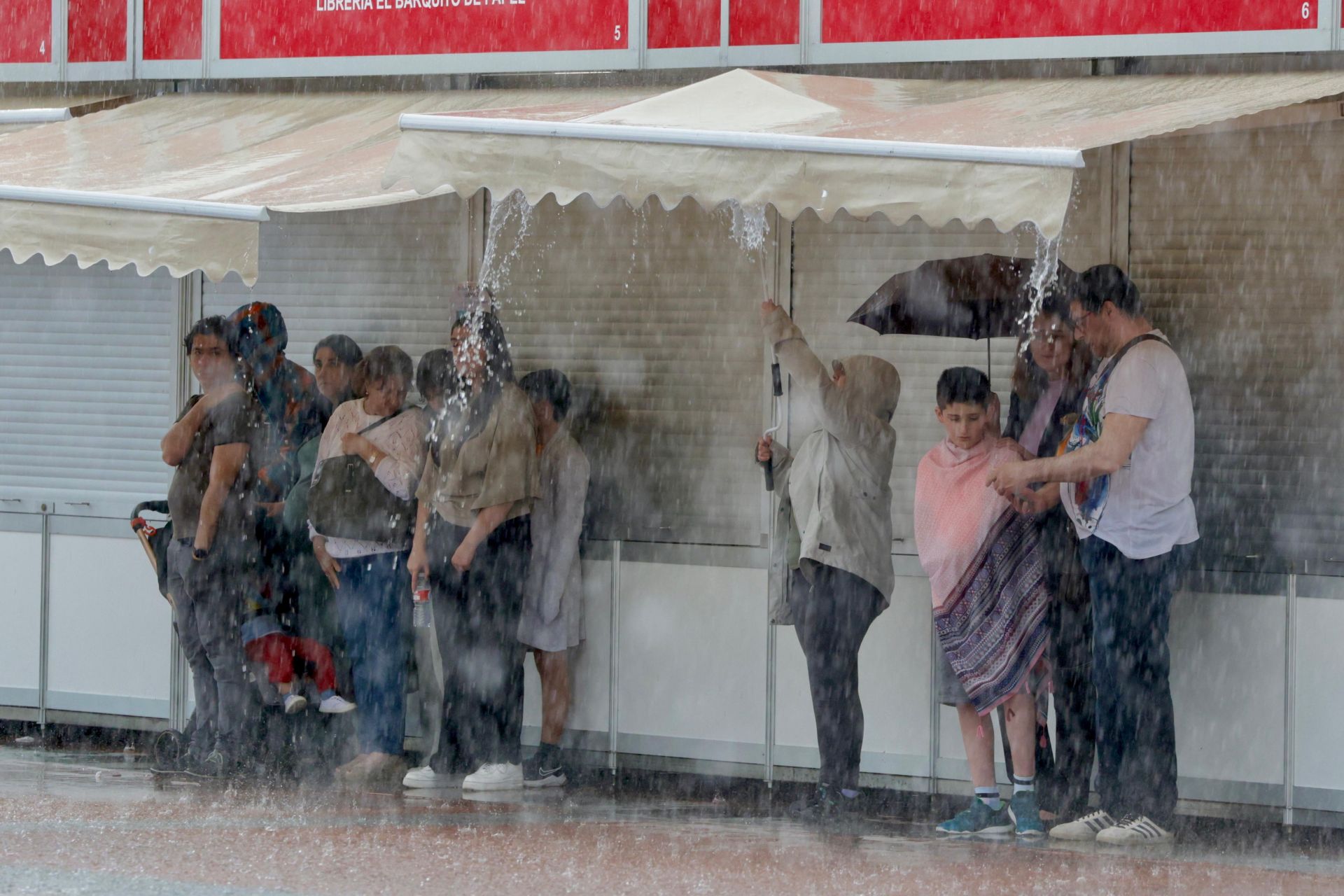Algunos ciudadanos se refugian de la lluvia bajo los toldos de las casetas de la Feria del Libro instalada en la Plaza Mayor.