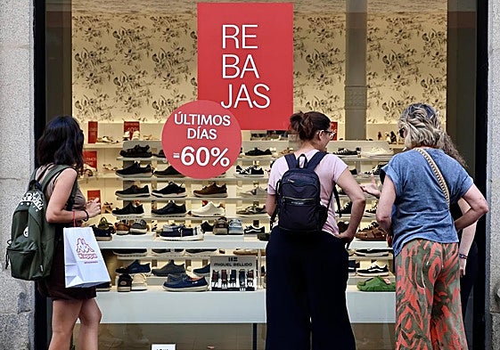 Viandantes se detienen a observar el escaparate de una zapatería de Valladolid que anuncia rebajas durante la campaña de verano.