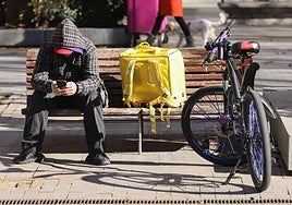 Un repartidor de Glovo espera que le entren pedidos móvil en mano en la Acera de Recoletos de Valladolid.