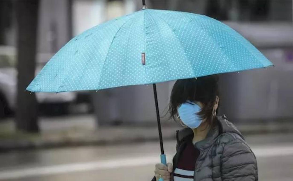Una mujer protegida con mascarilla y guantes se resguarda de la lluvia bajo su paragüas.