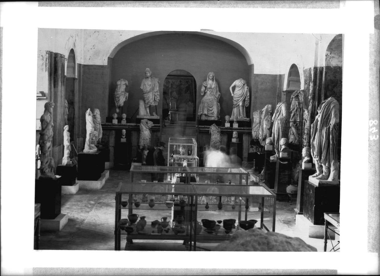 Sala de Santa Clara con visitantes. Nótese la excesiva abundancia de piezas expuestas, fruto de la incorporación de nuevos hallazgos (Anónimo, mediados de los años 50).. 