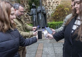 Varios cacereños muestran los 'cofraditos' en sus móviles, con el monumento de los cofrades al fondo.