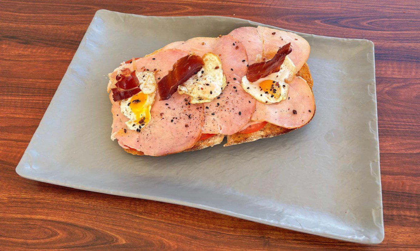 Se coge medio bollo de pan artesano, lo cubrimos de tomate en rodajas, ponemos una capa de queso semifundido, otra capa de lacón ahumado, colocamos encima tres huevos de codorniz a la plancha y unas lascas de jamón crujiente. Para terminar espolvoreamos topping de aguacate.