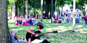 Una joven toca la guitarra en la Isla, entre un grupo de gente con mesas y sillas que conversan y juegan a las cartas. ::                             ANDY SOLÉ