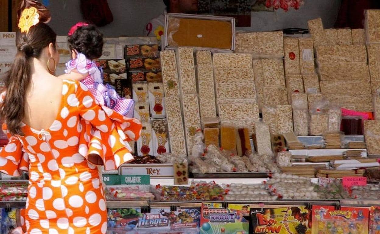 Una mujer con traje de flamenca, con una niña en brazos, frente a un puesto ambulantes de dulces y turrones. 