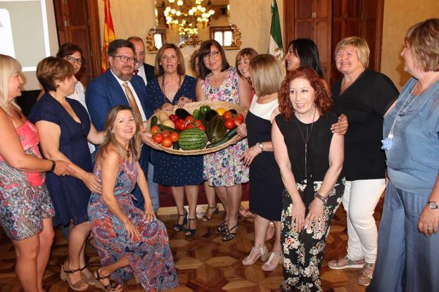 El consejero de Agricultura, acompañado por el delegado territorial, sostiene junto a mujeres integradas en la asociación una cesta con frutas y hortalizas de la tierra.