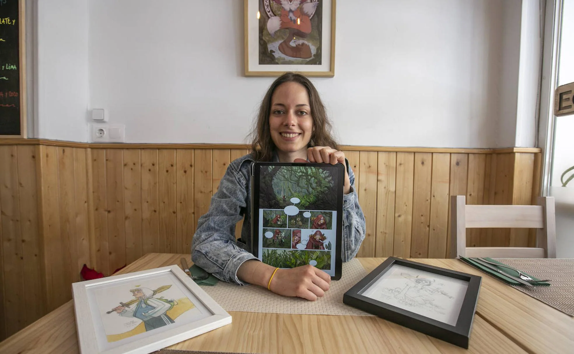 Maribel Conejero sostiene su tableta con uno de sus cómics, con algunas de las obras de la exposición- 