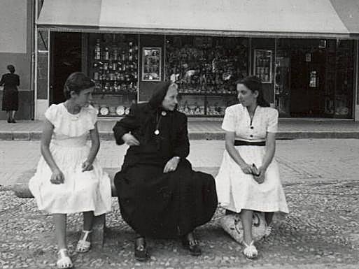 Una mujer mayor vestida de negro, sentada en medio de dos chicas jóvenes con vestidos blancos.