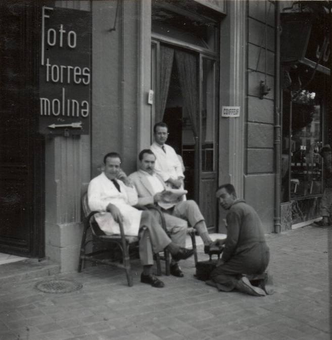 Un limpiabotas con un cliente. A cada lado, dos hombres que podrían ser peluqueros.