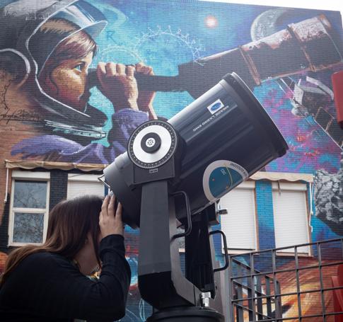 Una joven mira por un telescopio, delante del mural.