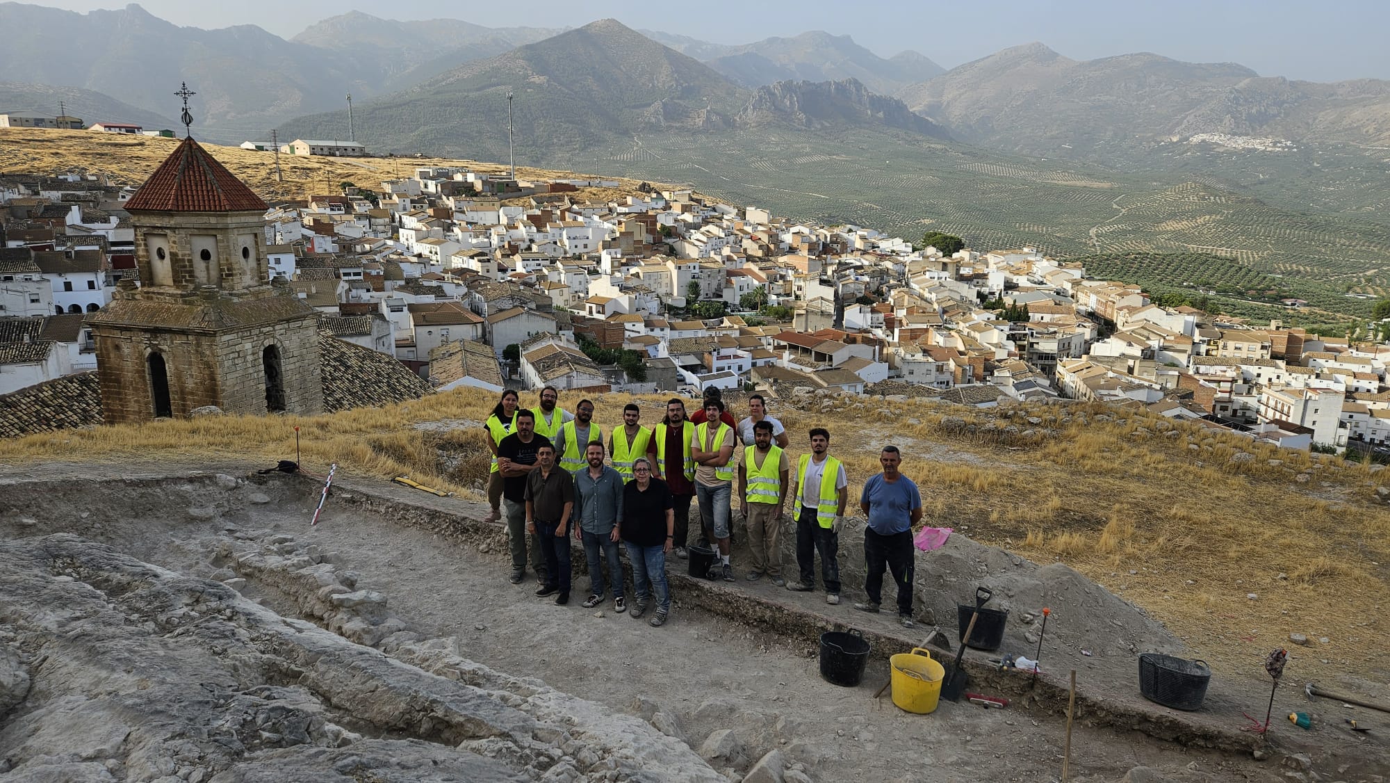 Zona de la excavación, el equipo de voluntarios con las autoridades y detrás una vista de Bedmar.