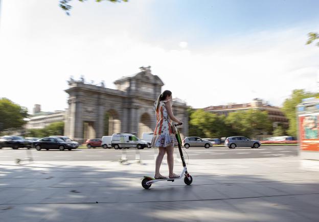Una mujer se desplaza en patinete eléctrico por una acera de Madrid frente a la Puerta de Alcalá. 