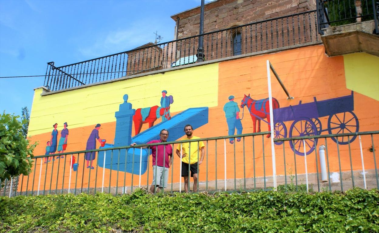 El alcalde y el pintor posan delante del mural que recuerda el antiguo uso de la fuente del pueblo. 