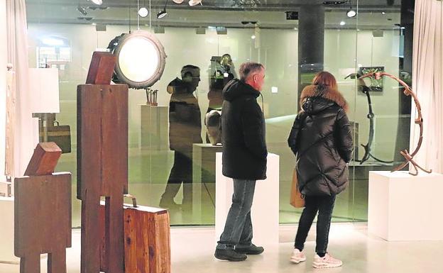 Imagen principal - Arriba, dos figuras de madera, portando una de ellas un maletín, observan el despliegue del resto de la exposición. Debajo, Una de las obras más llamativas de esta edición es un humanoide hecho de láminas de metal. 