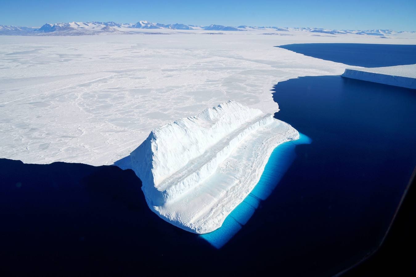 Imagen de la NASA publicada el 20 de diciembre de 2017 y tomada el 29 de noviembre de 2017 por la Operación IceBridge durante un vuelo a Victoria Land. La imagen muestra un iceberg flotando en el estrecho McMurdo de la Antártida. La parte del iceberg sumergida aparece más azul debido principalmente a la luz del agua. La parte inferior de algunos icebergs puede erosionarse y exponer hielo más viejo, más denso e increíblemente azul. La erosión puede cambiar la forma de un iceberg y hacer que se voltee, trayendo el hielo azul esculpido sobre la superficie del agua. La forma única en forma de escalón de este berg -comparado con el témpano tabular y más estable en la parte superior derecha de la imagen- sugiere que probablemente giró en algún momento. 