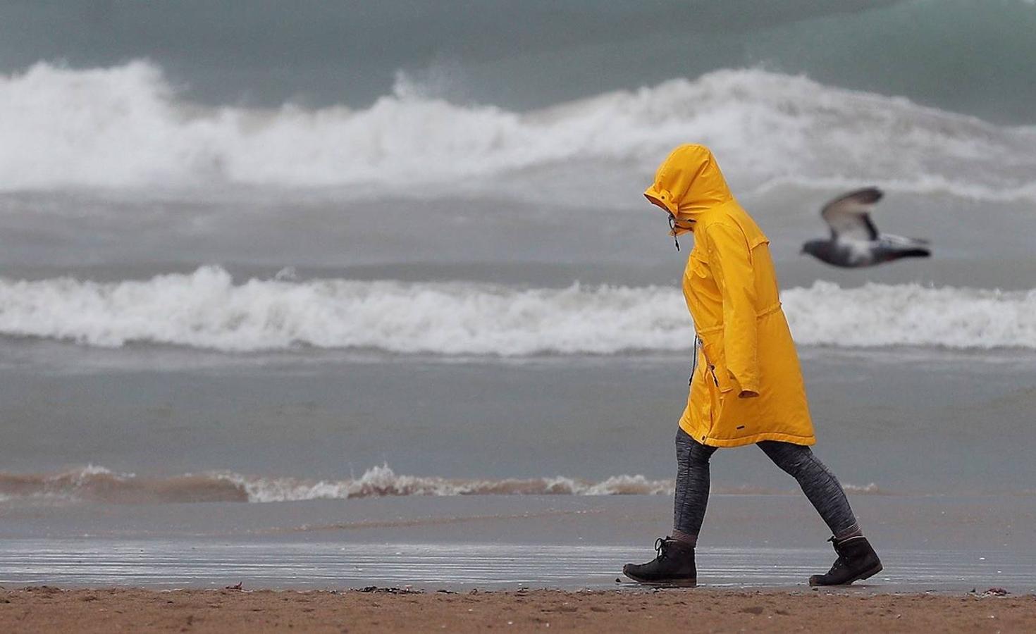 Una persona pasea por la playa de la Malvarrosa protegida contra la lluvia