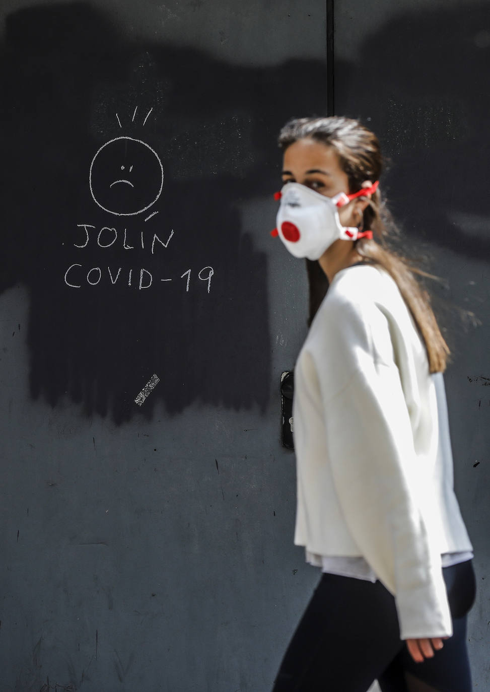 Una chica con mascarilla pasa junto a una pintada relacionada con el COVID-19