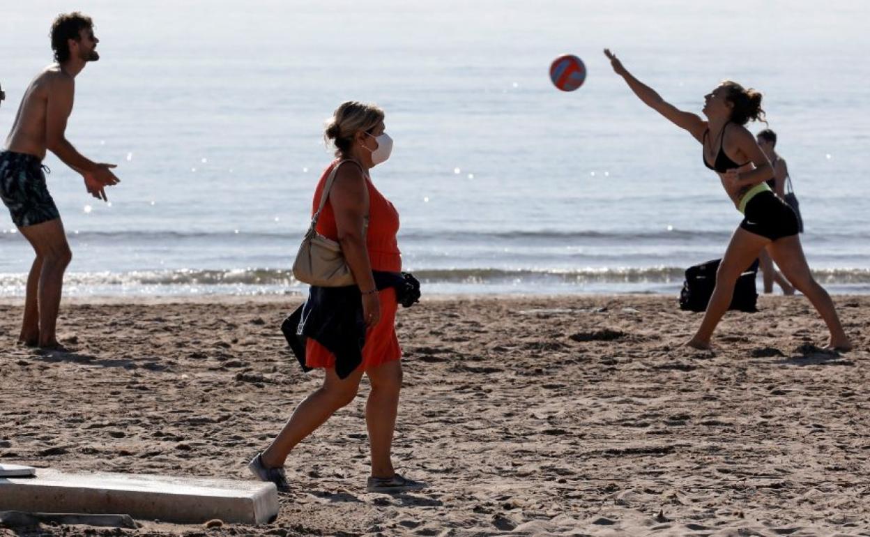 Una mujer con mascarilla en la playa ante dos jóvenes que juega con la pelota.