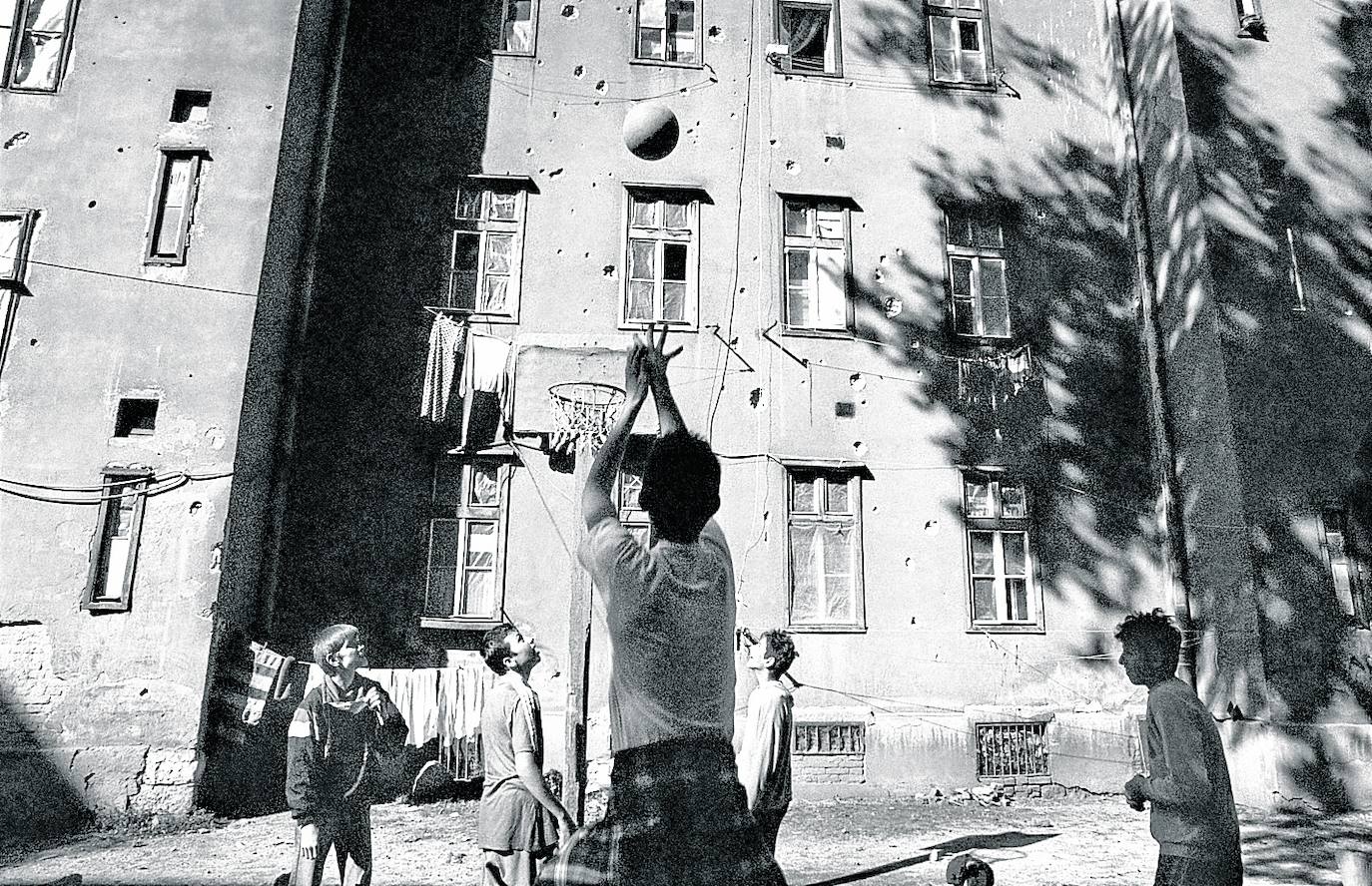 El chaval que intenta hacer una canasta, 25 años después frente a la misma casa acribillada en la siguiente foto.