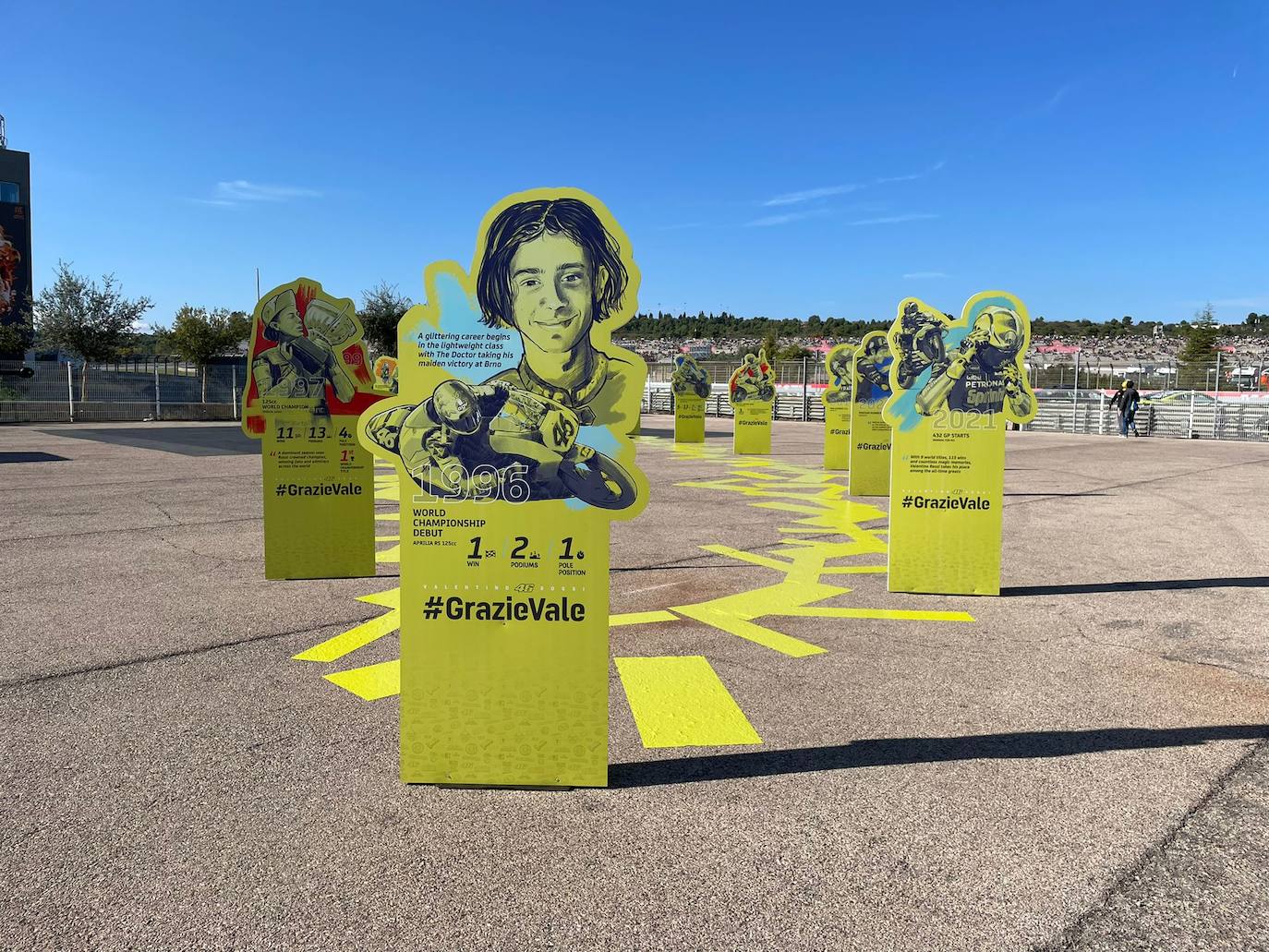 El paddock se ha teñido de amarillo este fin de semana. Como las gradas, que el sábado ya han expresado una muestra de intención de lo que será la carrera de este domingo. No es para nada exagerado afirmar que el 70% de los aficionados lucían merchandising de Valentino Rossi. Entre las muestras de homenaje que se le han dedicado por el momento, destaca la exposición de las nueve motos con las que ha sido campeón del mundo. Al lado está la 'Piazza Valentino Rossi', rotulada con una placa que simula ser de mármol y donde hay unos paneles amarilos que expresan los principales hitos del mito transalpino a lo largo de su carera. Más allá de esto, la mayoría de los camiones de los equipos tienen un panel, del mismo diseño, como muestra de gratitud al piloto por haber potenciado el Mundial de MotoGP durante sus más de 25 años de carrera. El propio piloto, en su mono, luce en la parte posterior este fin de semana un escueto pero representativo: 'Grazie'.