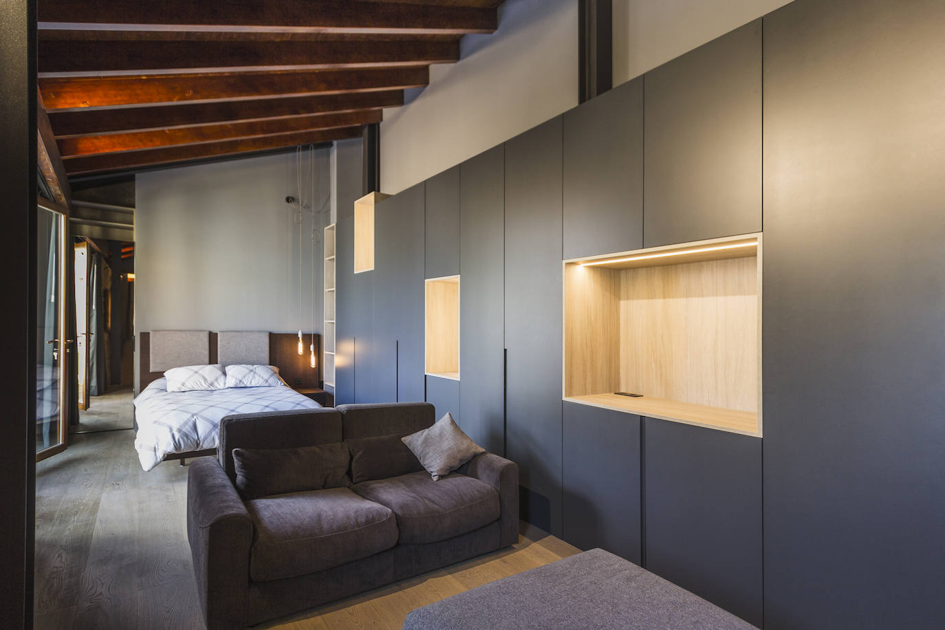 Habitación con cama y sofá de Grassoler y armariada lacada en gris con nichos de madera natural de roble.