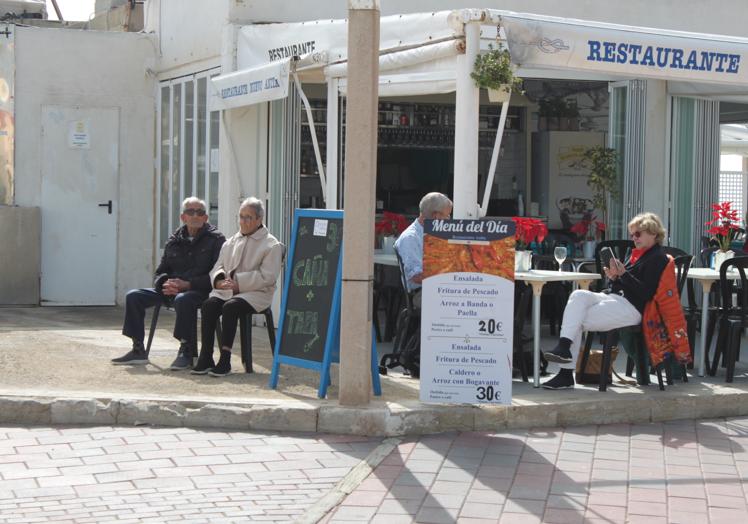 Dos tabarquinos disfrutan del sol mientras dos turistas se parapetan tras uno de los carteles de menú del día a 20 y 30 euros, de uno de los pocos bares que hay abiertos en invierno.