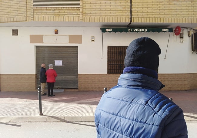 Varios vecinos miran el bar de la víctima, con el cartel que anuncia su fallecimiento en la puerta, este sábado, en la calle García Berlanga de la localidad.