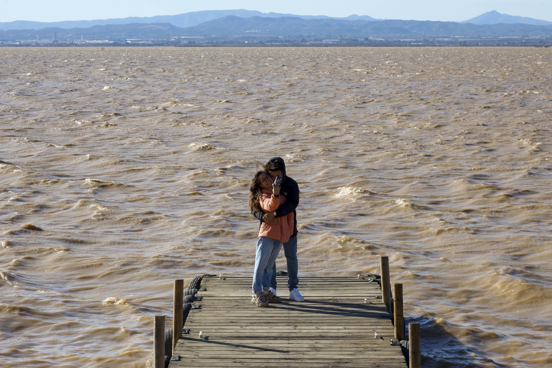 Dos personas en el embarcadero del lago, cuya agua presenta un tono marrón.