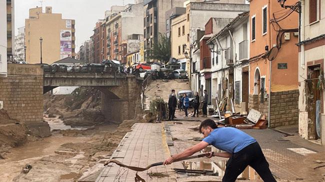 Imagen antes - Alejandro, un vecino de Chiva, era con su pala un David contra Goliat el día después de la dana. Al fondo, la pila de coches arrastrados por el agua. En la calle ha abierto ya la tienda de ropa de Cunquero, la barbería de Richi, el locutorio y el quiosco de la esquina.