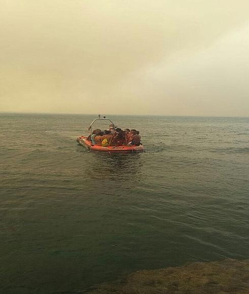 Una barca de rescate de Cruz Roja, llena de bañistas, con el resplandor del fuego de fondo.