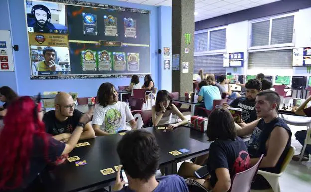 Un grupo de jóvenes disfruta de una partida a un juego de cartas mientras se proyecta un enfentamiento en un videojuego 'online'. 