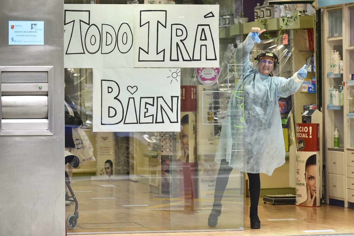 20-03-2020. Ventanas y escaparates cuelgan carteles con el lema ‘Todo irá bien’, en muchos casos decorado con el arco iris. La población trata de reforzar así el ánimo durante el duro confinamiento. En la imagen, una farmacéutica de la calle García Alix de Murcia reparte apoyo moral.