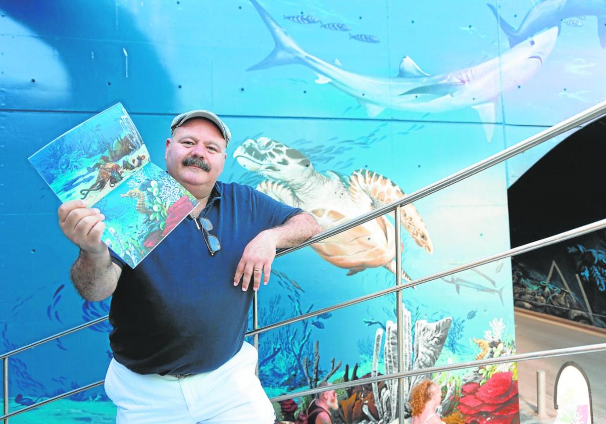 El artista con el catálogo de fotografías de Pablo Sánchez en la mano y un fondo azul protagonizado por una tortuga, un caballito de mar y más seres marinos.