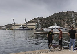 Tres jóvenes en el cantil del puerto y enfrente el 'Ysabel' y dos yates atracados en el muelle del astillero de Navantia.
