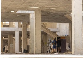 Dos hombres entran a un habitación construida bajo unas escaleras, mientras al fondo se ve otra chabola con ropa y varios muebles.