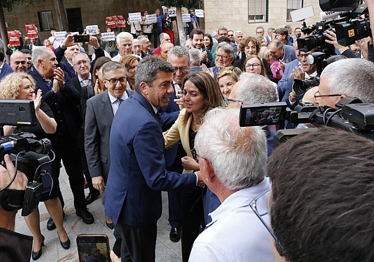 Mazón, ayer en Orihuela, recibe aplausos de dirigentes del PP. Al fondo, los manifestantes que pidieron su dimisión.