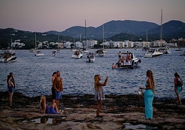 Un grupo de jóvenes se hace fotos frente al mar en Ibiza, donde hay fondeados varios barcos abarrotados.