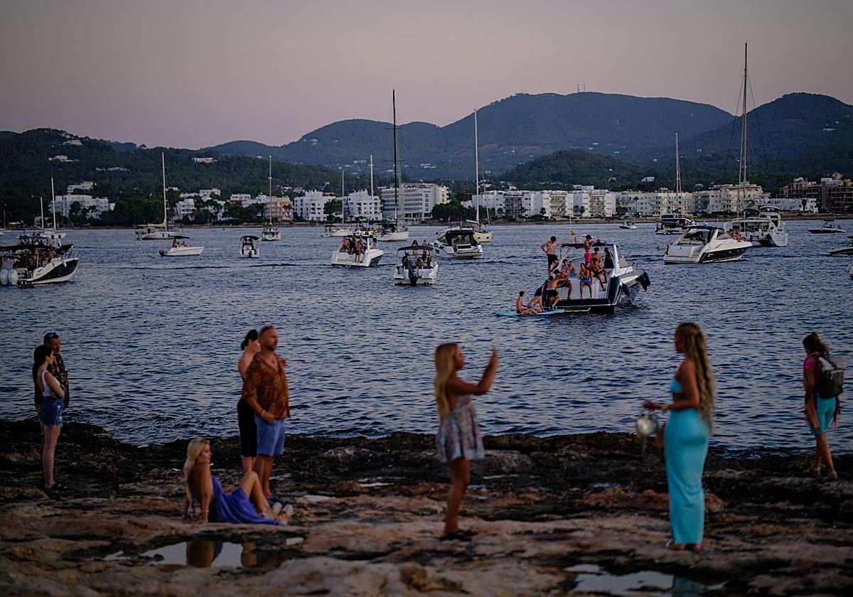 Un grupo de jóvenes se hace fotos frente al mar en Ibiza, donde hay fondeados varios barcos abarrotados.