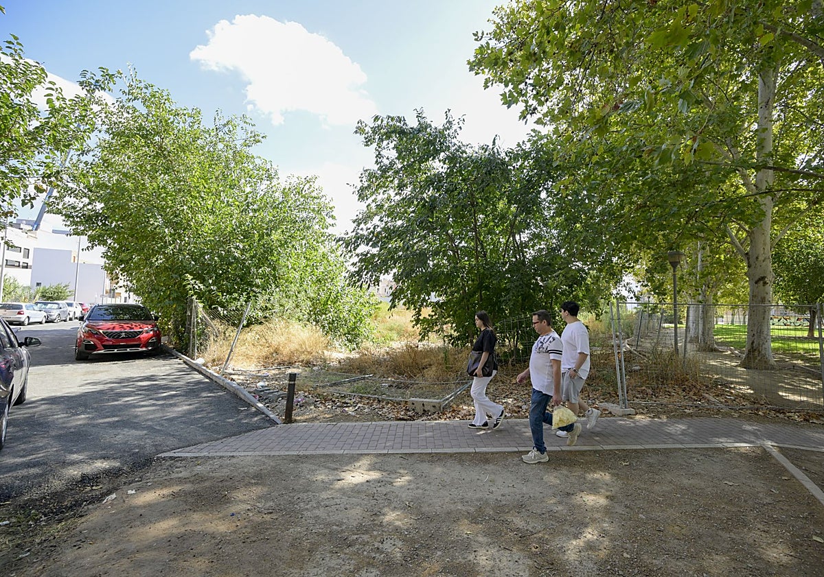 Imagen principal - En la imagen superior, varios transeúntes pasan por delante de un tramo forzado de la valla perimetral. Abajo, detalle una zona central de la zona verde, con muestras de haber sido usada para pernoctar al raso. En la fotografía inferior, un vecino señala las casas vecinas cercanas a un entorno cubierto de malezas y con restos de palés de madera rotos.