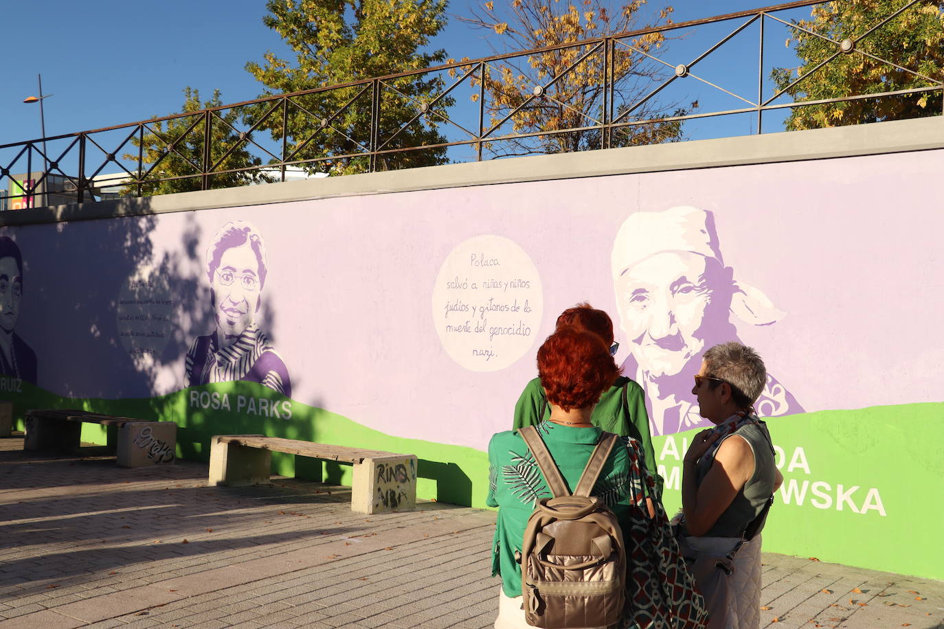 Tres mujeres conversan con el mural de las mujeres pioneras en la historia al fondo.