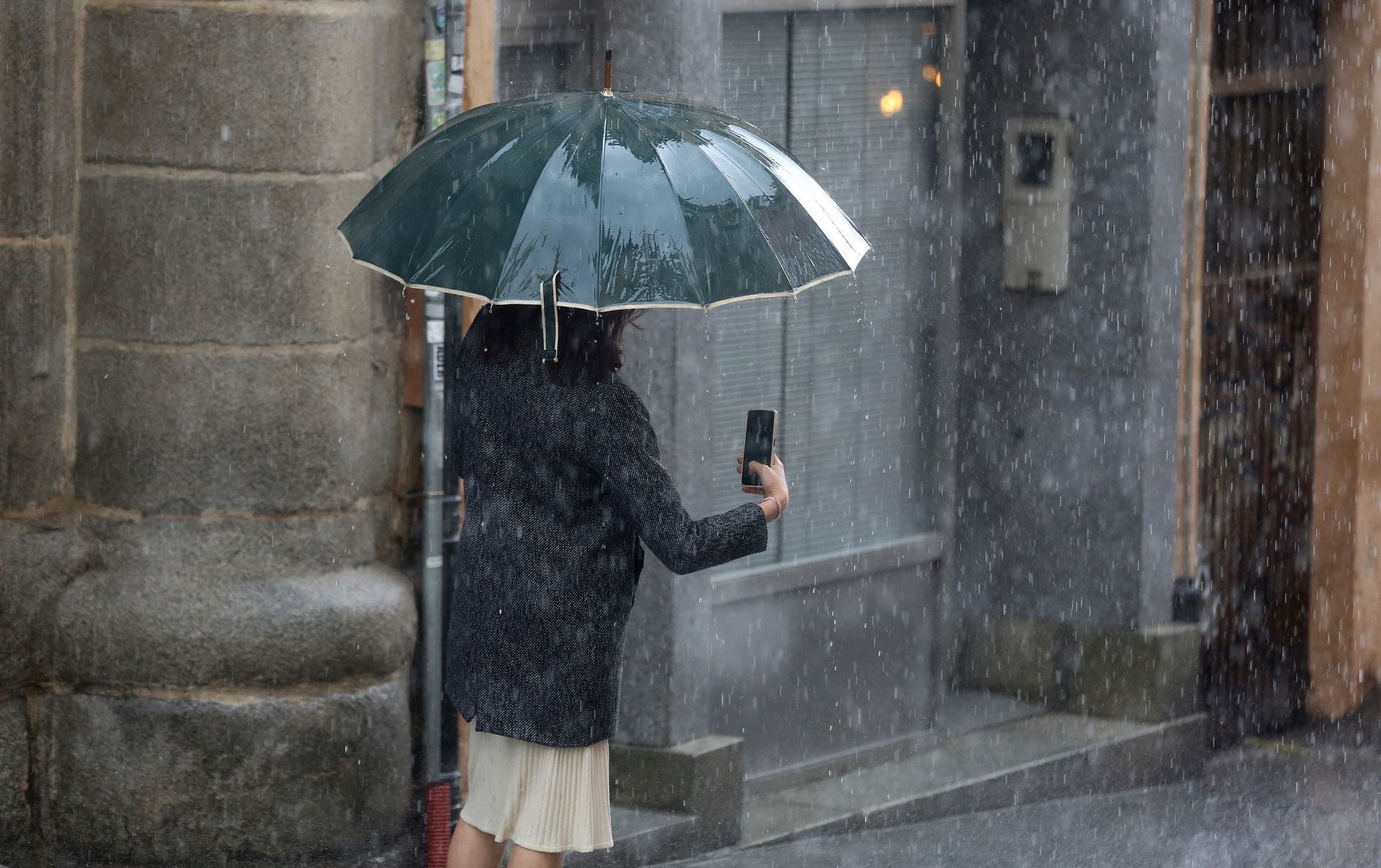 Una mujer camina bajo la lluvia con el paraguas abierto y mirando el teléfono en una imagen de archivo.